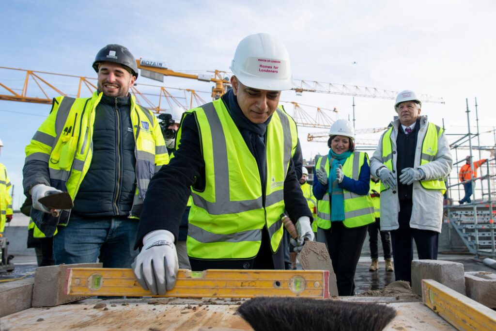 Sadiq Khan uses a spirit level on a construction site where they're building homes. He's wearing a bright yellow high vis jacket.