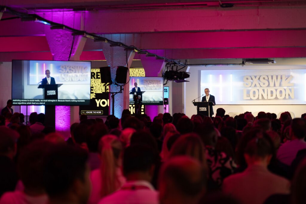 Sadiq Khan speaks in front of a large audience in London. The lighting is purple and blue.